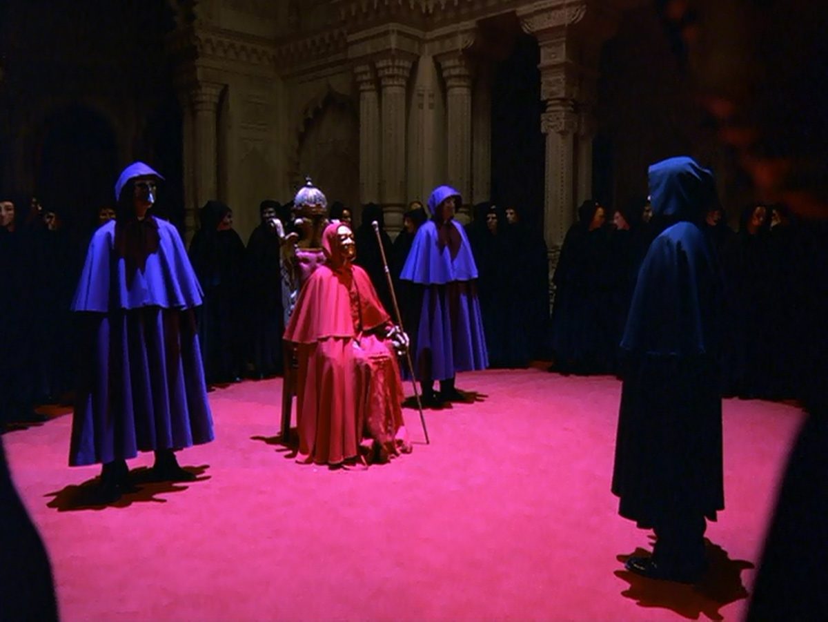 The masked ceremony: a ring of cloaked figures around a seated figure in red, on a patterned floor